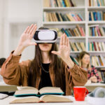 Young Caucasian brunette girl student working with VR simulator in library. Young woman in casual wear and virtual reality glasses sitting at desk and touching air. VR goggles for studying concept