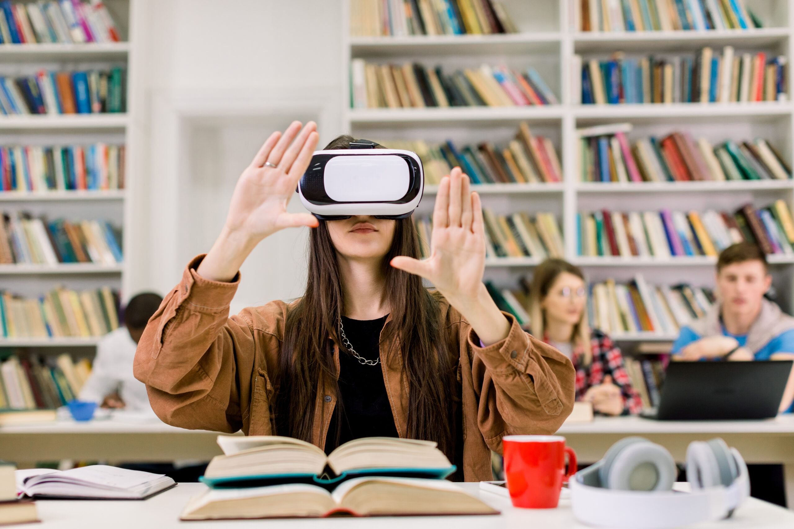 Young Caucasian brunette girl student working with VR simulator in library. Young woman in casual wear and virtual reality glasses sitting at desk and touching air. VR goggles for studying concept